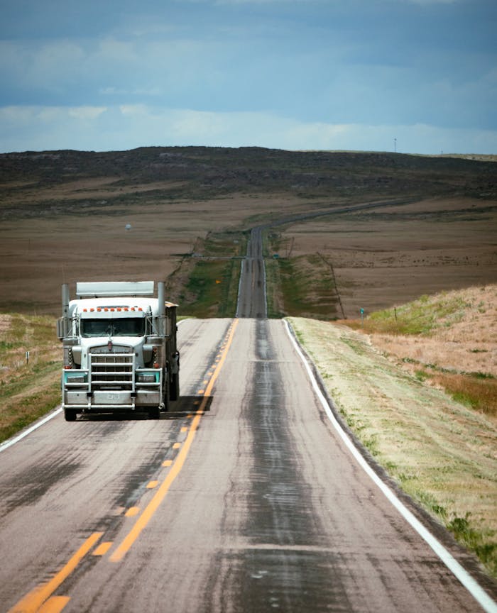 A truck traveling on a long empty highway through expansive countryside under a blue sky.